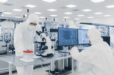 Pharma and MedTech industry: two employees in white protecting suits working in a laboratory on computer and on a microscope in a biological or pharmaceutical setting